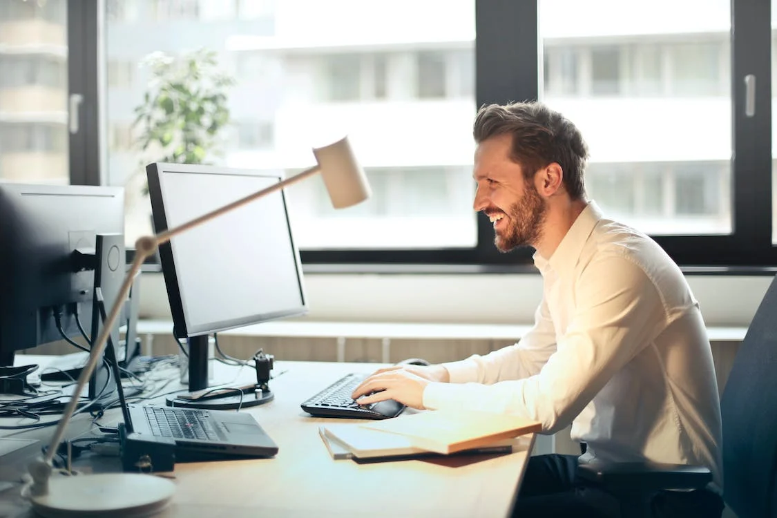 leende man i vit skjorta som sitter vid ett skrivbord och tittar på sin datorskärm